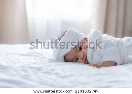 Adorable baby girl in white bathrobe lie prone looking down with sleepy emotion on white bed in bed room, blurred white curtain background and light in morning.