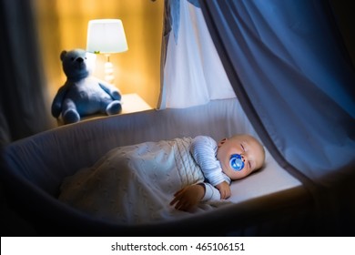 Adorable Baby With Pacifier Sleeping In Bassinet With Canopy At Night. Little Boy In Pajamas Taking A Nap In Dark Room With Crib, Lamp And Toy Bear. Bed Time For Kids. Bedroom And Nursery Interior.