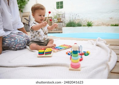 Adorable baby playing with colorful educational toys on a blanket by the poolside. Parent sitting next to the baby. Bright and cheerful outdoor setting ideal for childhood development and bonding. - Powered by Shutterstock