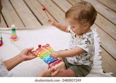 An adorable baby playing a colorful xylophone with the support of a parent, enjoying playful and musical moments outdoors on a cozy blanket. - Powered by Shutterstock