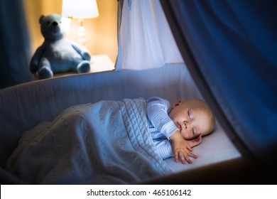 Adorable Baby Sleeping In Blue Bassinet With Canopy At Night. Little Boy In Pajamas Taking A Nap In Dark Room With Crib, Lamp And Toy Bear. Bed Time For Kids. Bedroom And Nursery Interior.