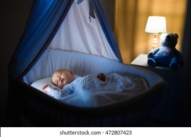 Adorable Baby Sleeping In Blue Bassinet With Canopy At Night. Little Boy In Pajamas Taking A Nap In Dark Room With Crib, Lamp And Toy Bear. Bed Time For Kids. Bedroom And Nursery Interior.