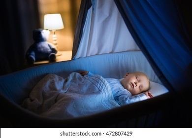 Adorable Baby Sleeping In Blue Bassinet With Canopy At Night. Little Boy In Pajamas Taking A Nap In Dark Room With Crib, Lamp And Toy Bear. Bed Time For Kids. Bedroom And Nursery Interior.