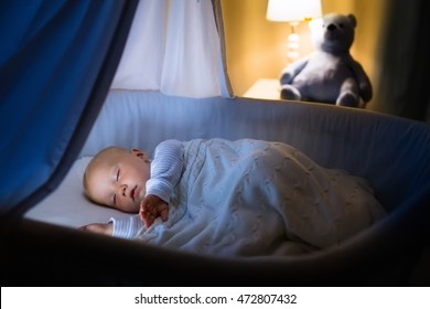 Adorable Baby Sleeping In Blue Bassinet With Canopy At Night. Little Boy In Pajamas Taking A Nap In Dark Room With Crib, Lamp And Toy Bear. Bed Time For Kids. Bedroom And Nursery Interior.