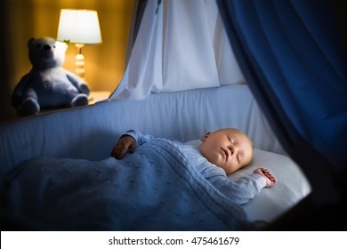 Adorable Baby Sleeping In Blue Bassinet With Canopy At Night. Little Boy In Pajamas Taking A Nap In Dark Room With Crib, Lamp And Toy Bear. Bed Time For Kids. Bedroom And Nursery Interior.