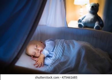 Adorable Baby Sleeping In Blue Bassinet With Canopy At Night. Little Boy In Pajamas Taking A Nap In Dark Room With Crib, Lamp And Toy Bear. Bed Time For Kids. Bedroom And Nursery Interior.