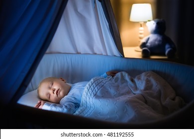 Adorable Baby Sleeping In Blue Bassinet With Canopy At Night. Little Boy In Pajamas Taking A Nap In Dark Room With Crib, Lamp And Toy Bear. Bed Time For Kids. Bedroom And Nursery Interior.