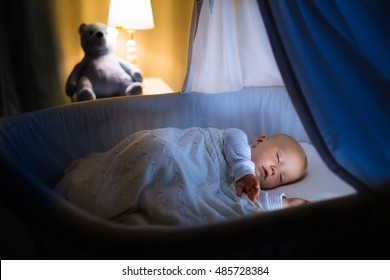 Adorable baby sleeping in blue bassinet with canopy at night. Little boy in pajamas taking a nap in dark room with crib, lamp and toy bear. Bed time for kids. Bedroom and nursery interior.
