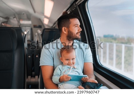 The adorable baby smiles while holding the phone in her hand and sitting on her father's lap as they ride the train