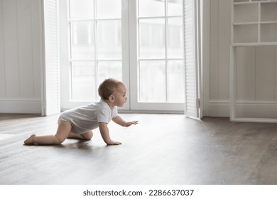 Adorable baby wearing white bodysuit, crawling on knees on floor at home. Curious active little infant child learning to move on warm heating safe floor, passing by window in background - Shutterstock ID 2286637037
