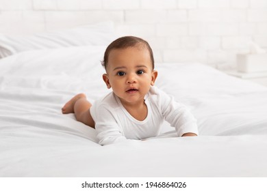 Adorable Black Baby Toddler Crawling On Bed Smiling To Camera Posing In Bedroom At Home. Portrait Of Cute Little Newborn Kid Lying On Stomach. Child Care And Happy Childhood. Full Length