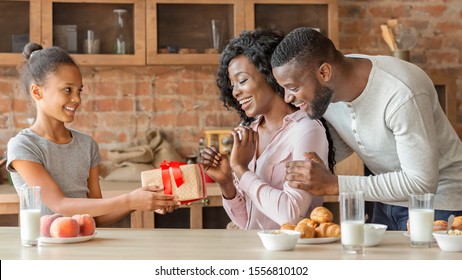 Adorable Black Girl Holding Birthday Gift For Her Surprised Mom, Daddy Standing Behind, Kitchen Interior, Panorama With Copy Space