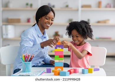 Adorable black kid curly little girl and child development specialist attractive young woman playing with colorful wooden bricks, sitting at table and making pyramid together, smiling