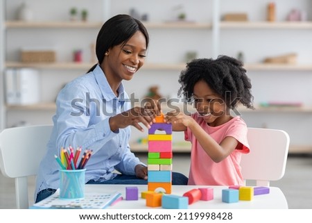 Adorable black kid curly little girl and child development specialist attractive young woman playing with colorful wooden bricks, sitting at table and making pyramid together, smiling