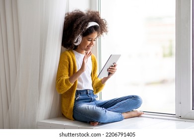 Adorable Black Kid Having Video Call With Friends, Cute African American Preteen Girl Sitting On Windowsill At Home, Using Wireless Headphones, Waving At Digital Tablet Screen, Copy Space