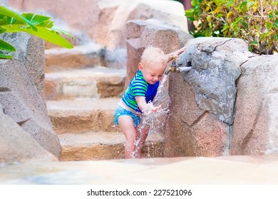 Adorable blond curly baby boy in a sun protection shirt and swimming diaper playing with water tap at pool side on a hot summer day