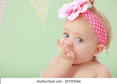 Adorable blond hair big blue eyed baby girl with pink flower elastic head band on head and pony tail in hair sucking eating and tasting vanilla sponge cake on green background with flag bunting behind