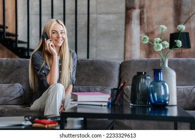 Adorable blonde Swedish young woman talking by phone looks at camera sitting on couch with vases and books toothy smiles at home. Successful female realtor talks with client from luxury house. - Shutterstock ID 2233329613