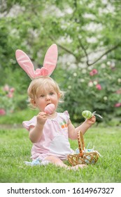 Adorable blonde toddler girl wearing bunny ears playing with Easter eggs in a basket sitting in a sunny garden with first pink spring flowersKid in bunny ears and rabbit costume.  - Shutterstock ID 1614967327