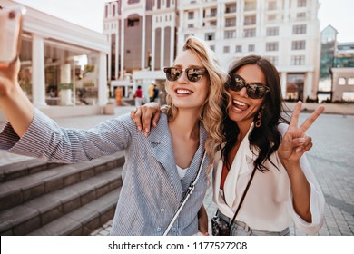 Adorable Blonde Woman With Curly Hair Making Selfie On The Street. Carefree Female Friends Posing With Peace Sign On City Background.