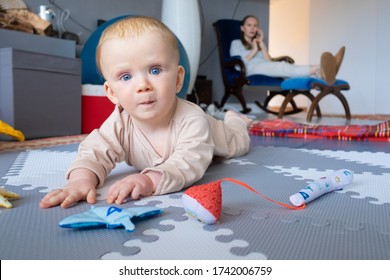 Adorable Blue Eyed Baby Posing While Playing On Floor, Trying To Crawl, Looking At Camera With Wide Eyes. Busy Mom Talking On Phone In Background. Childhood Or Staying At Home Concept