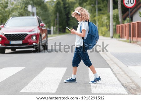 Adorable boy with a backpack, headphones and cellphone goes through the pedestrian crossing, not looking at red cars let him through