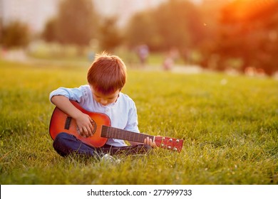 Adorable Boy With Guitar, Sitting On The Grass On Sunset