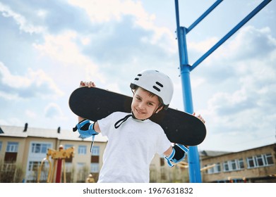 Adorable Boy In A Protective Helmet And Equipment Posing With A Skateboard On His Shoulders Behind His Head Against The Background Of A Clear Sky