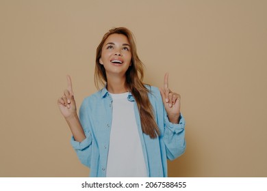 Adorable Brunette Female With Long Straight Hair Wears Casual White Tshirt And Blue Shirt, Indicates Upwards On Blank Copy Space, Notices Something On Ceiling, Stands Against Beige Wall