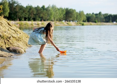 Adorable brunette kid girl playing with paper boat at the lake in the evening sunlight
