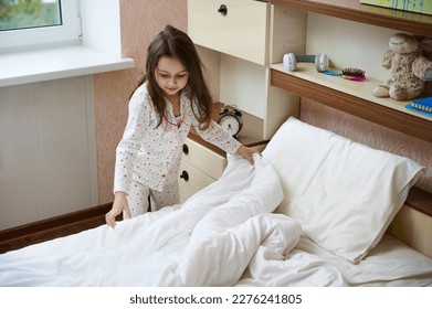 Adorable Caucasian 5 years old child, a lovely little girl in pajamas, straightens bed sheets and duvet on her bed after sleeping, cleans her light sunny bedroom in the morning. Child's daily routine - Shutterstock ID 2276241805