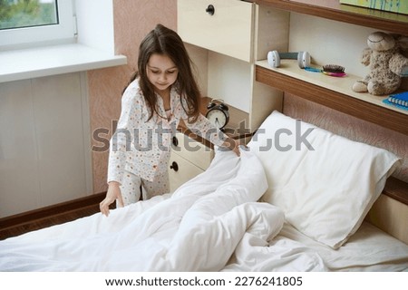Adorable Caucasian 5 years old child, a lovely little girl in pajamas, straightens bed sheets and duvet on her bed after sleeping, cleans her light sunny bedroom in the morning. Child's daily routine