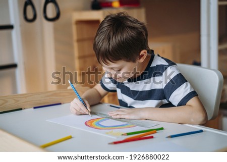adorable caucasian boy of elementary age drawing a rainbow with pencils sitting at the desk in his room at home. Image with selective focus