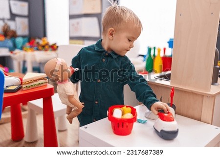 Adorable caucasian boy standing with relaxed expression playing with baby doll at kindergarten