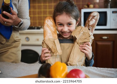 Adorable Caucasian Child, Funny Little Girl With Two Loaves Of Whole Grain Sourdough Bread, Smiling With A Cheerful Toothy Smile, Looking At Camera, Standing At Kitchen Table, Unpacking Grocery Bag