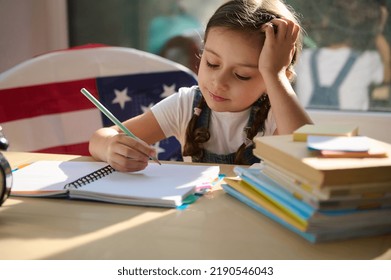 Adorable Caucasian Child, Inspired Schoolgirl With Two Pigtails In White T-shirt And Casual Denim, Learning The Writing On First Grade, Doing Homework While Sitting On A Chair With American Flag