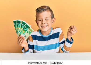 Adorable Caucasian Kid Holding Israel 50 Shekels Sitting On The Table Screaming Proud, Celebrating Victory And Success Very Excited With Raised Arm 