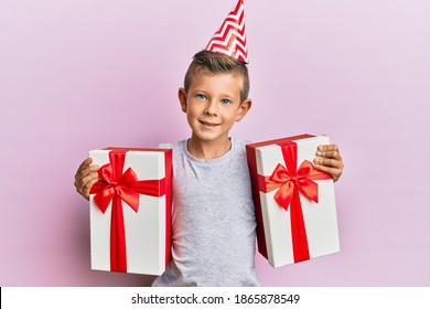 Adorable Caucasian Kid Wearing Birthday Hat Holding Presents Smiling With A Happy And Cool Smile On Face. Showing Teeth. 