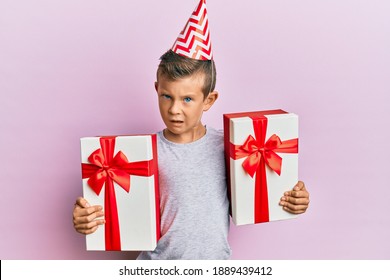 Adorable Caucasian Kid Wearing Birthday Hat Holding Presents In Shock Face, Looking Skeptical And Sarcastic, Surprised With Open Mouth 