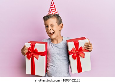 Adorable Caucasian Kid Wearing Birthday Hat Holding Presents Winking Looking At The Camera With Sexy Expression, Cheerful And Happy Face. 