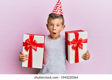 Adorable Caucasian Kid Wearing Birthday Hat Holding Presents Afraid And Shocked With Surprise And Amazed Expression, Fear And Excited Face. 