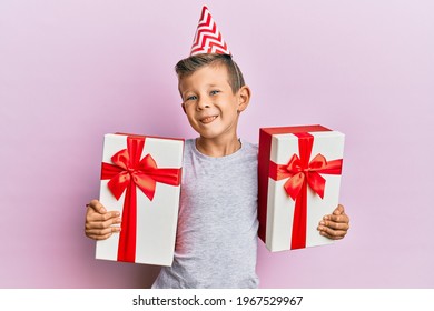 Adorable Caucasian Kid Wearing Birthday Hat Holding Presents Sticking Tongue Out Happy With Funny Expression. 