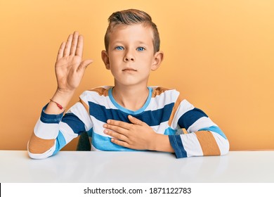 Adorable Caucasian Kid Wearing Casual Clothes Sitting On The Table Swearing With Hand On Chest And Open Palm, Making A Loyalty Promise Oath 