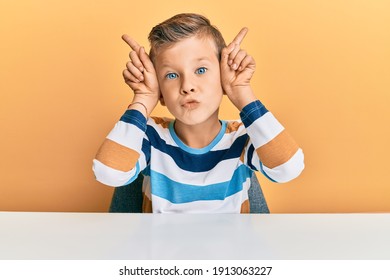 Adorable Caucasian Kid Wearing Casual Clothes Sitting On The Table Doing Funny Gesture With Finger Over Head As Bull Horns 