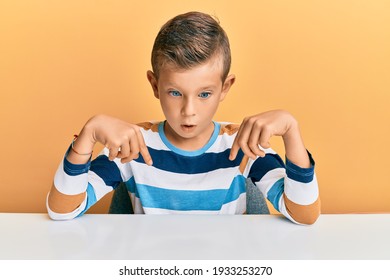 Adorable Caucasian Kid Wearing Casual Clothes Sitting On The Table Pointing Down With Fingers Showing Advertisement, Surprised Face And Open Mouth 