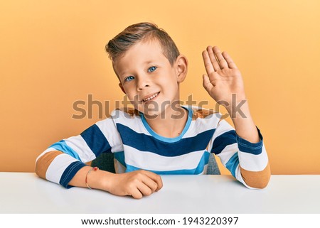 Adorable caucasian kid wearing casual clothes sitting on the table waiving saying hello happy and smiling, friendly welcome gesture 