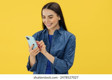 Adorable caucasian woman in casual clothes standing with a smartphone in her hand while buying online or typing message isolated over yellow background.