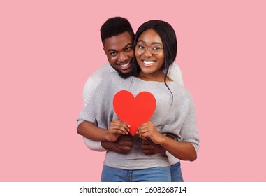 Adorable cheerful african man and woman celebrating St Valentine's Day together, holding red heart, pink background