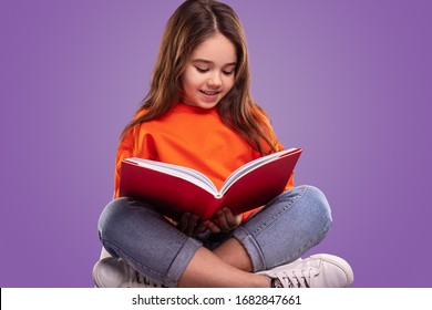 Adorable Child In Casual Outfit Smiling And Reading Interesting Book While Sitting Crossed Legged Against Violet Background