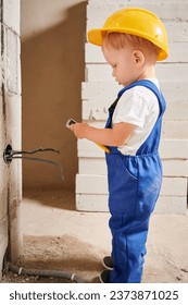 Adorable child construction worker using wire stripper cutter tool while installing electric cables and socket in wall. Kid in safety helmet mounting electrical wiring in apartment under renovation. - Shutterstock ID 2373871025